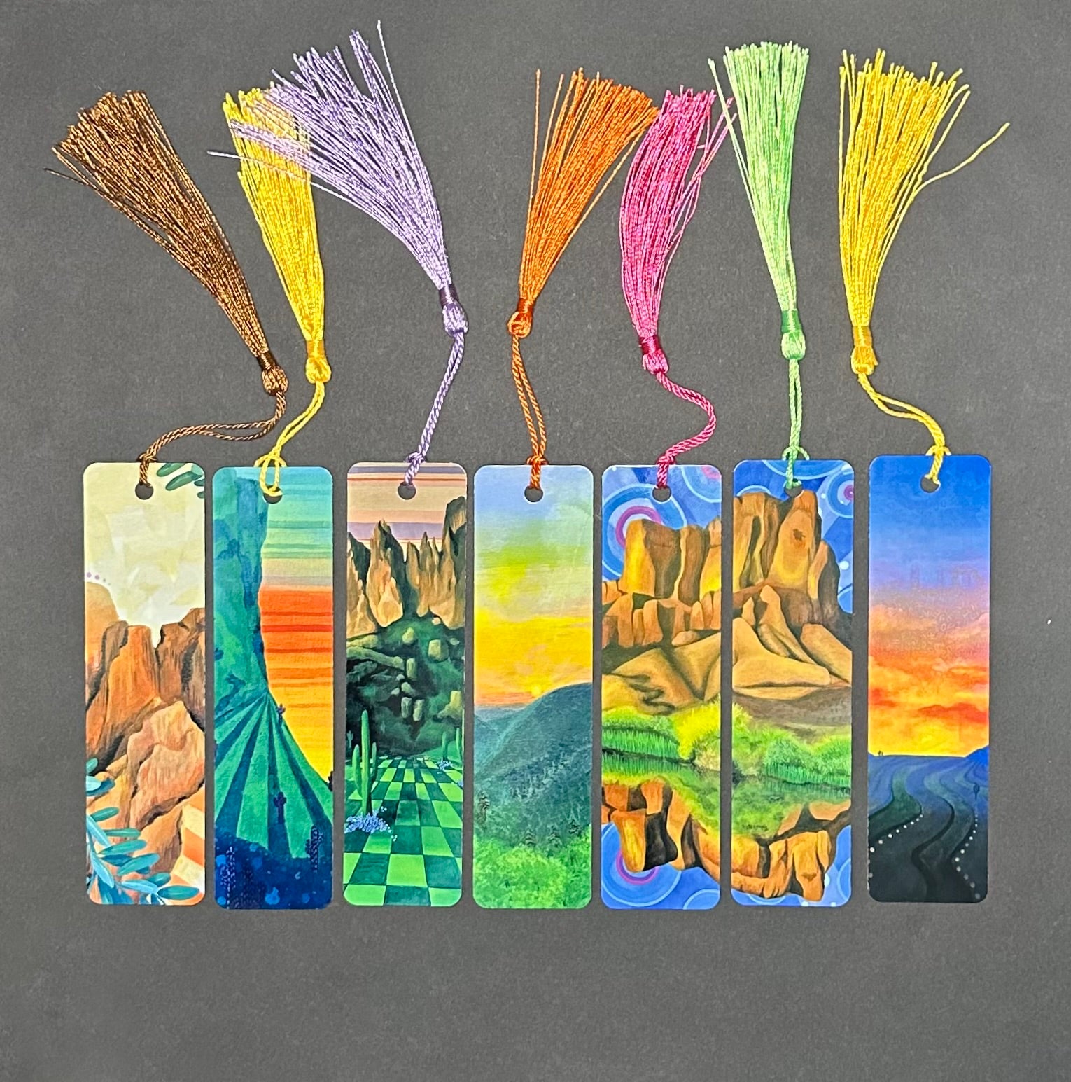 Metal bookmarks with colorful art prints featuring various colored tassels in a straight line on a black background.